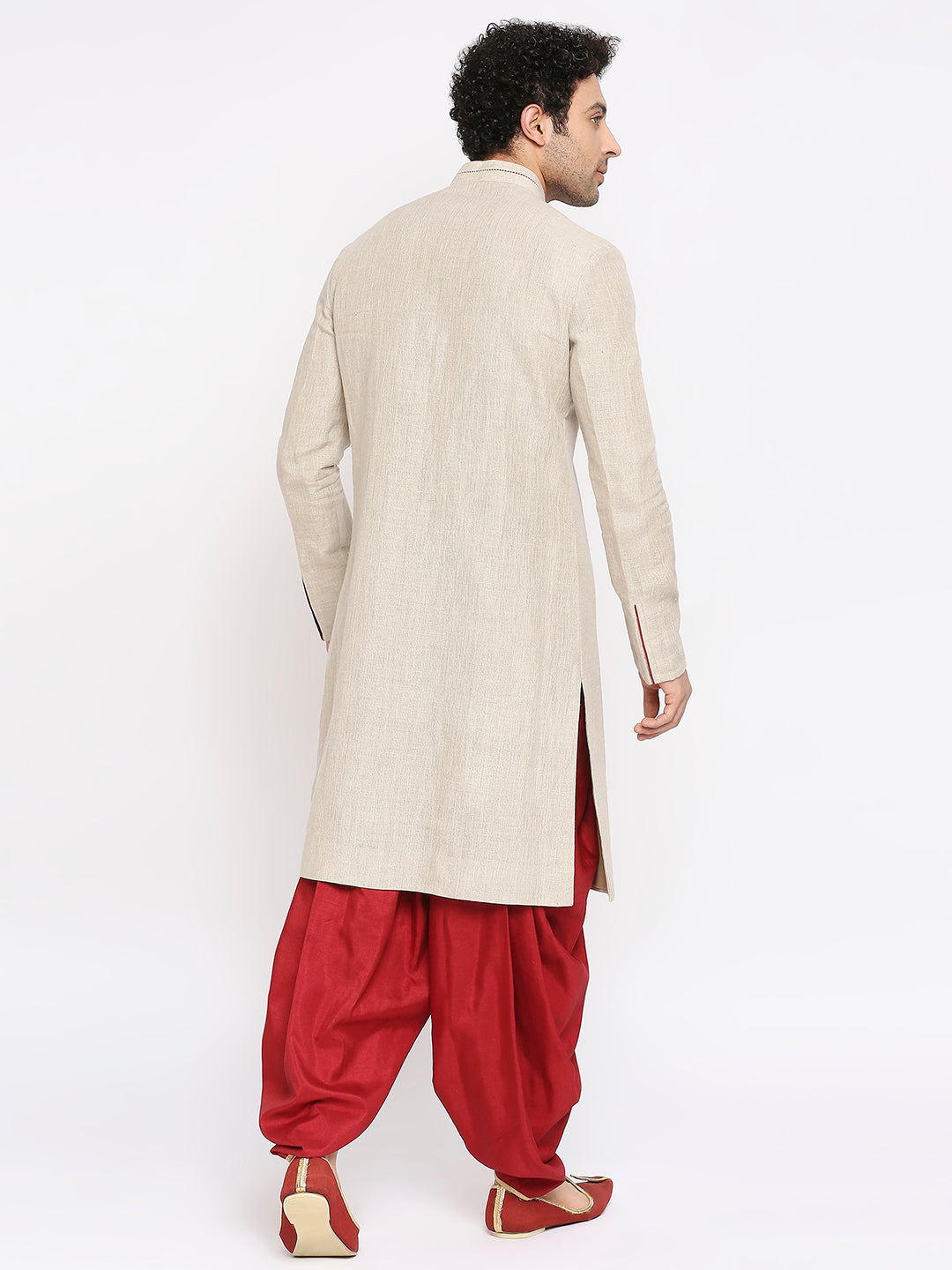A man shown from the back wearing a light beige linen kurta over red sharara pants and brown shoes, photographed against a plain white studio background.