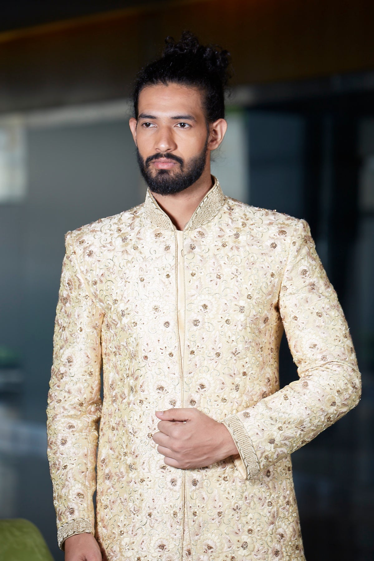 A man wearing a cream-colored sherwani jacket with intricate gold and brown floral embroidery, standing against a blurred dark background.