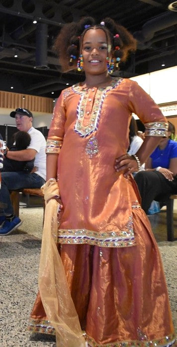 A girl modeling a traditional South Asian outfit in a metallic copper-orange color with intricate silver and gold embroidery, standing in an indoor event space.