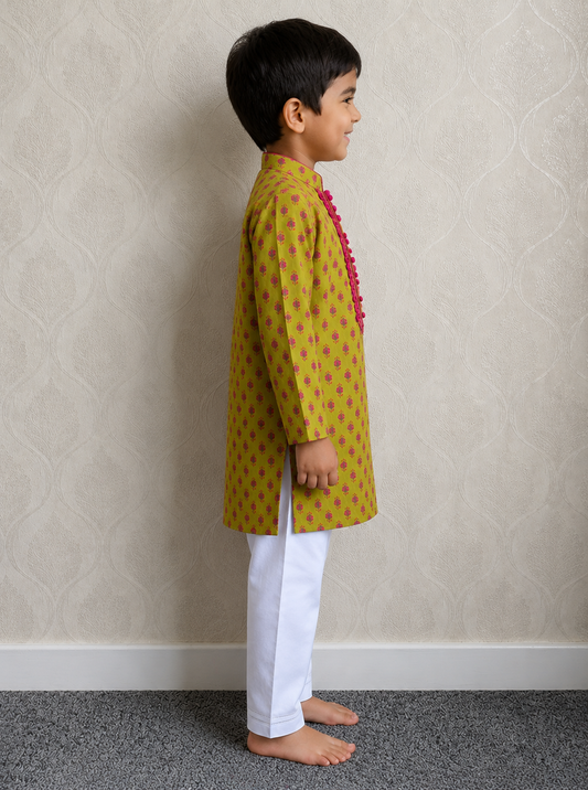 A young boy standing in profile wearing a green kurta with small pink motifs and a pink tassel detail, paired with white trousers, against a textured beige wall.