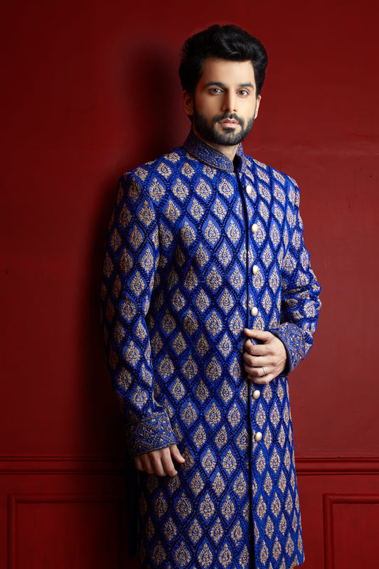 A man wearing a royal blue sherwani jacket with intricate gold and silver geometric patterns, standing against a dark red paneled wall.