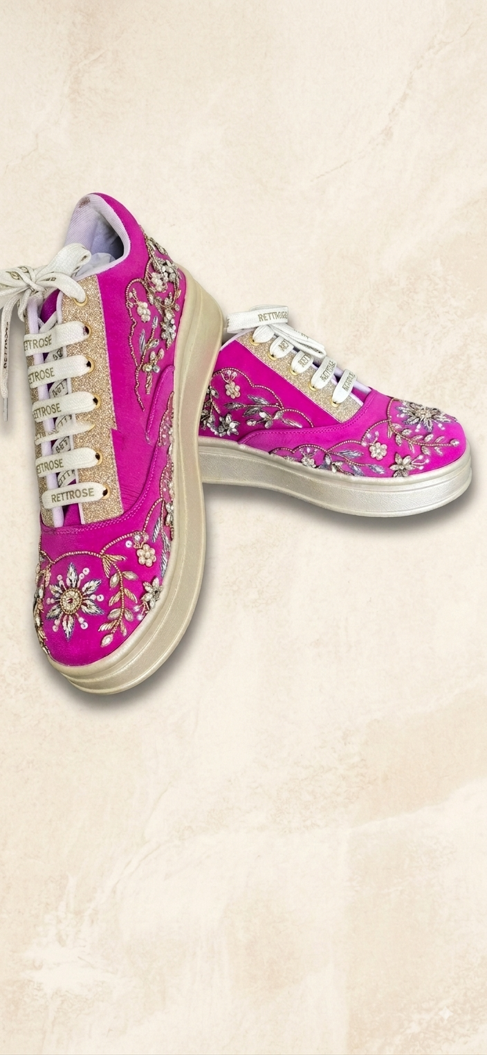 A pair of low-top sneakers in vibrant pink with white laces, white soles, and detailed silver and gold floral embroidery on the sides, displayed against a textured beige background.