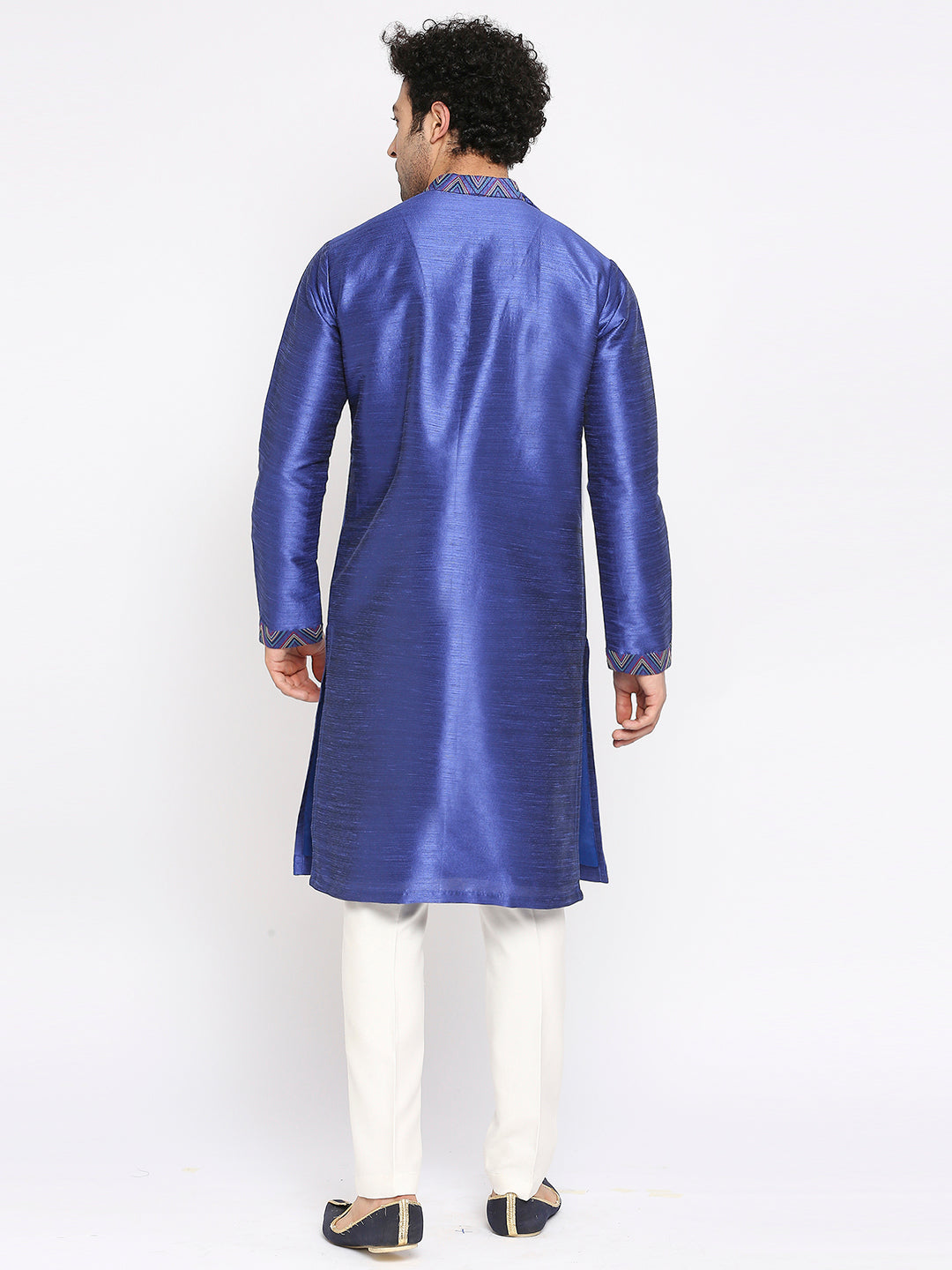 A man viewed from the back wearing a royal blue satin kurta with white trousers and black sandals, set against a plain white studio background.