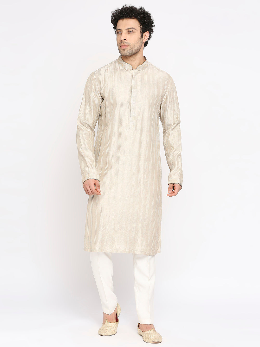 A man modeling a light beige long-sleeve kurta with subtle vertical stripes, paired with white trousers and beige shoes, against a plain white studio background.