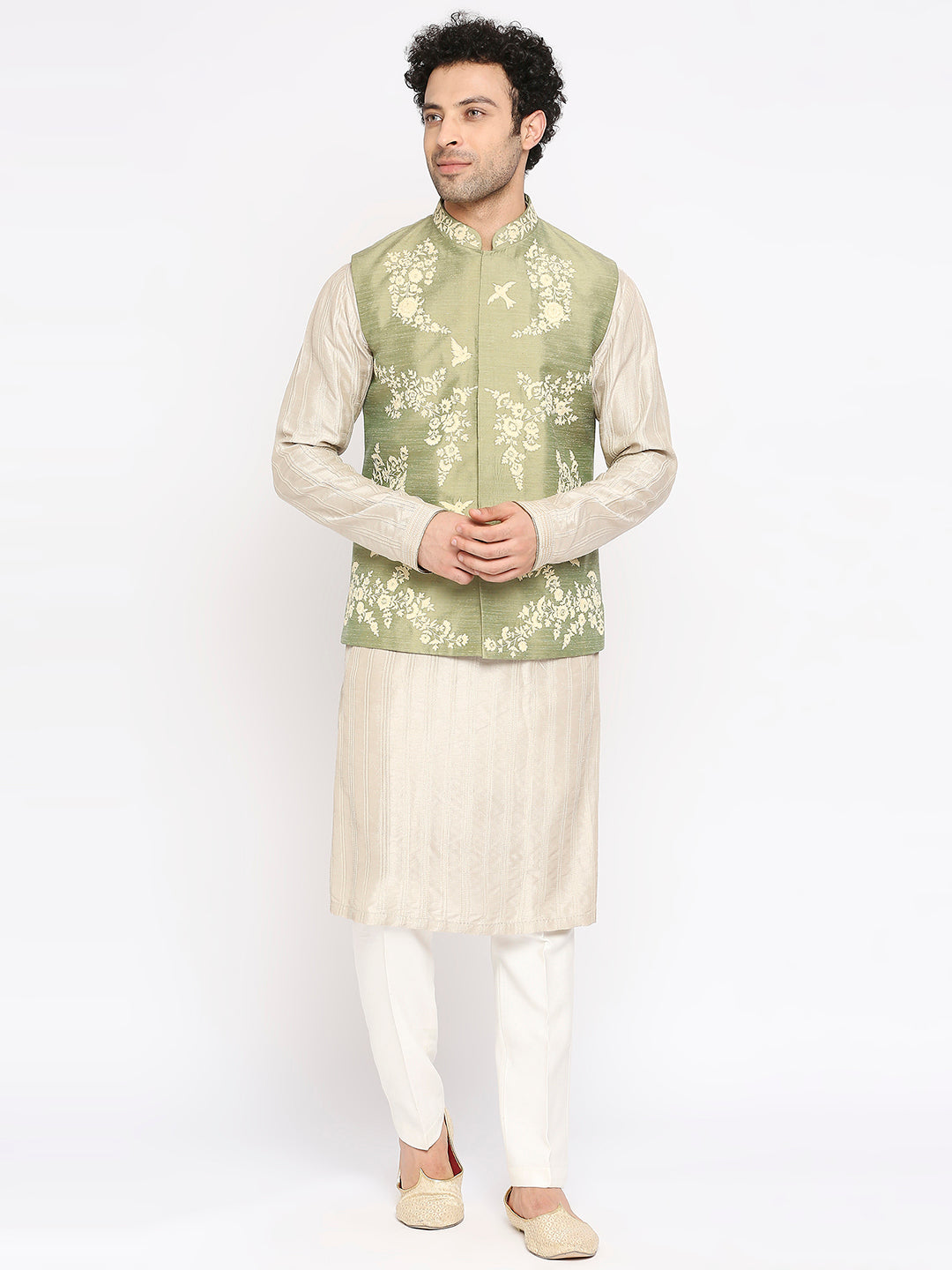 A man modeling a traditional Indian outfit consisting of a light sage green embroidered waistcoat over a cream kurta, paired with white trousers and beige loafers, against a plain white studio background.