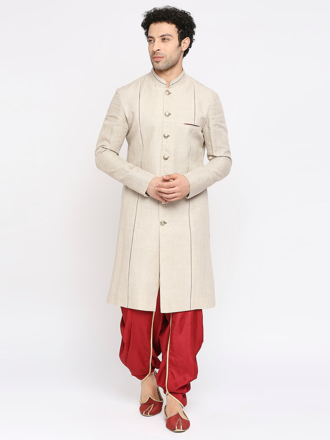 A man modeling a traditional beige sherwani jacket with gold buttons, paired with red pants and red shoes, against a plain white studio background.