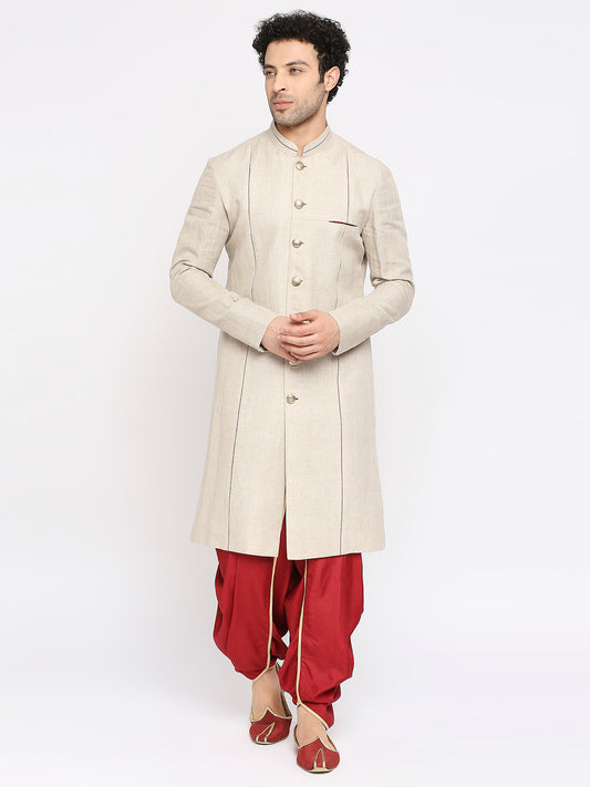 A man modeling a traditional beige sherwani jacket with gold buttons, paired with red pants and red shoes, against a plain white studio background.