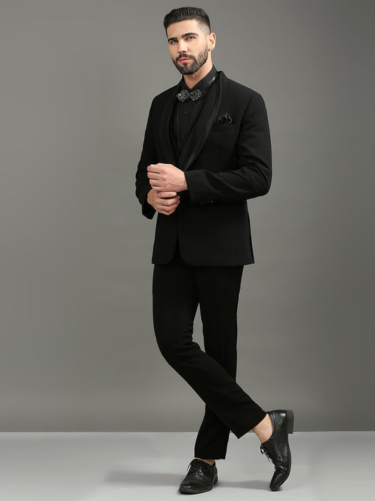 A man modeling a black formal suit with a black tuxedo jacket, black trousers, a black shirt, and black dress shoes against a solid gray studio background.