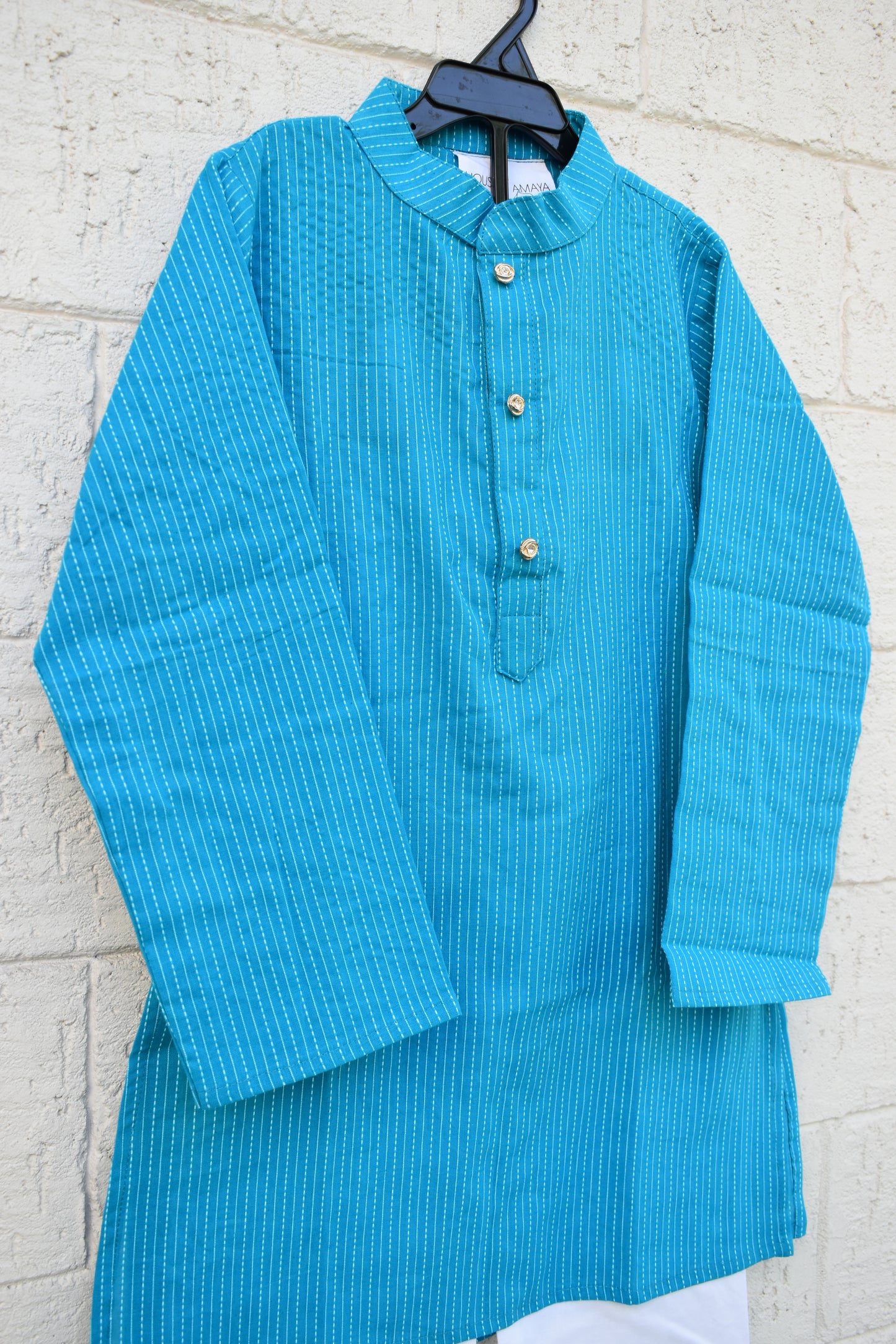A bright turquoise blue textured long-sleeve tunic top with a mandarin collar and three gold buttons, hanging on a black hanger against a white painted brick wall.