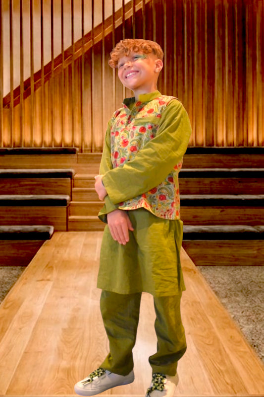 A smiling young boy with curly light brown hair models a traditional Indian outfit consisting of an olive green kurta and trousers with a red and green floral patterned vest, standing on a wooden runway in front of a wooden staircase.
