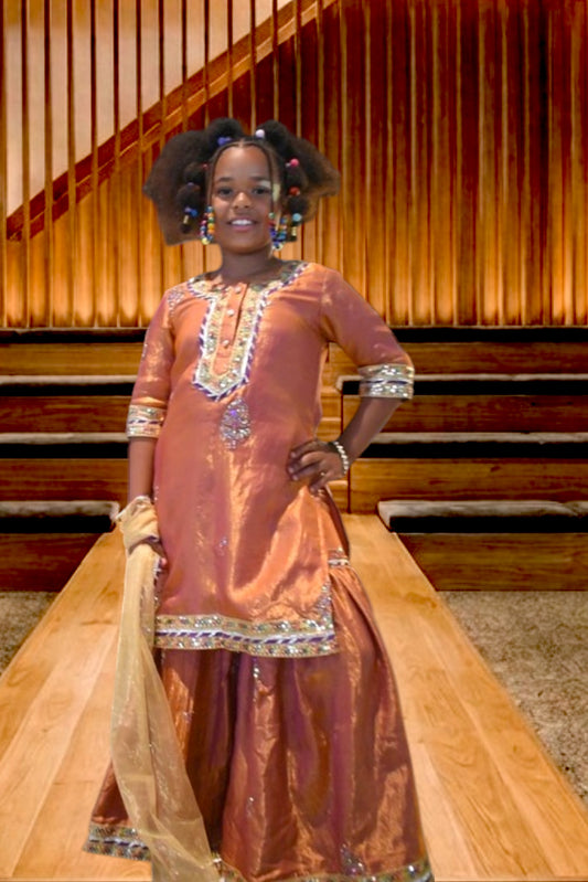 A girl modeling a traditional metallic copper-brown salwar kameez outfit with silver embroidery, standing on a wooden runway in front of a wooden slatted wall.