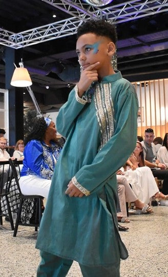 A child model walks the runway wearing a teal green traditional long kurta with silver embroidery down the front and cuffs, set against a fashion show audience background.