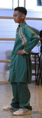 A child model wearing a teal green traditional outfit with silver embroidery on the chest, standing in an indoor setting.