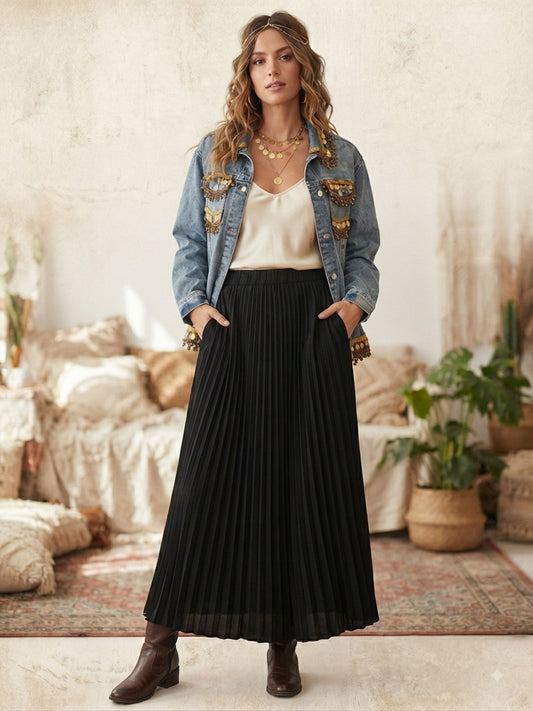 A woman stands in a styled indoor room wearing a long black pleated midi skirt with side pockets, layered under a light blue denim jacket with gold embroidery, a cream camisole, and brown ankle boots.