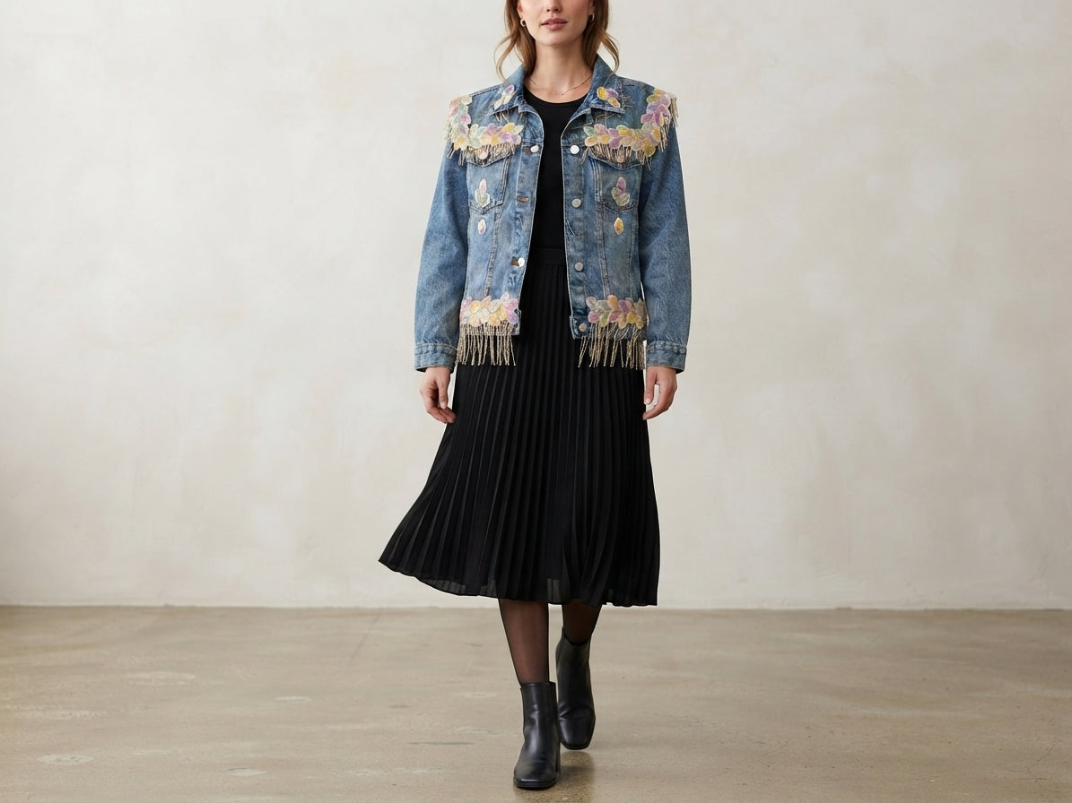 A model wearing a light blue denim jacket with colorful floral embroidery on the shoulders and sleeves, paired with a black pleated midi skirt and black ankle boots, standing against a neutral beige wall.