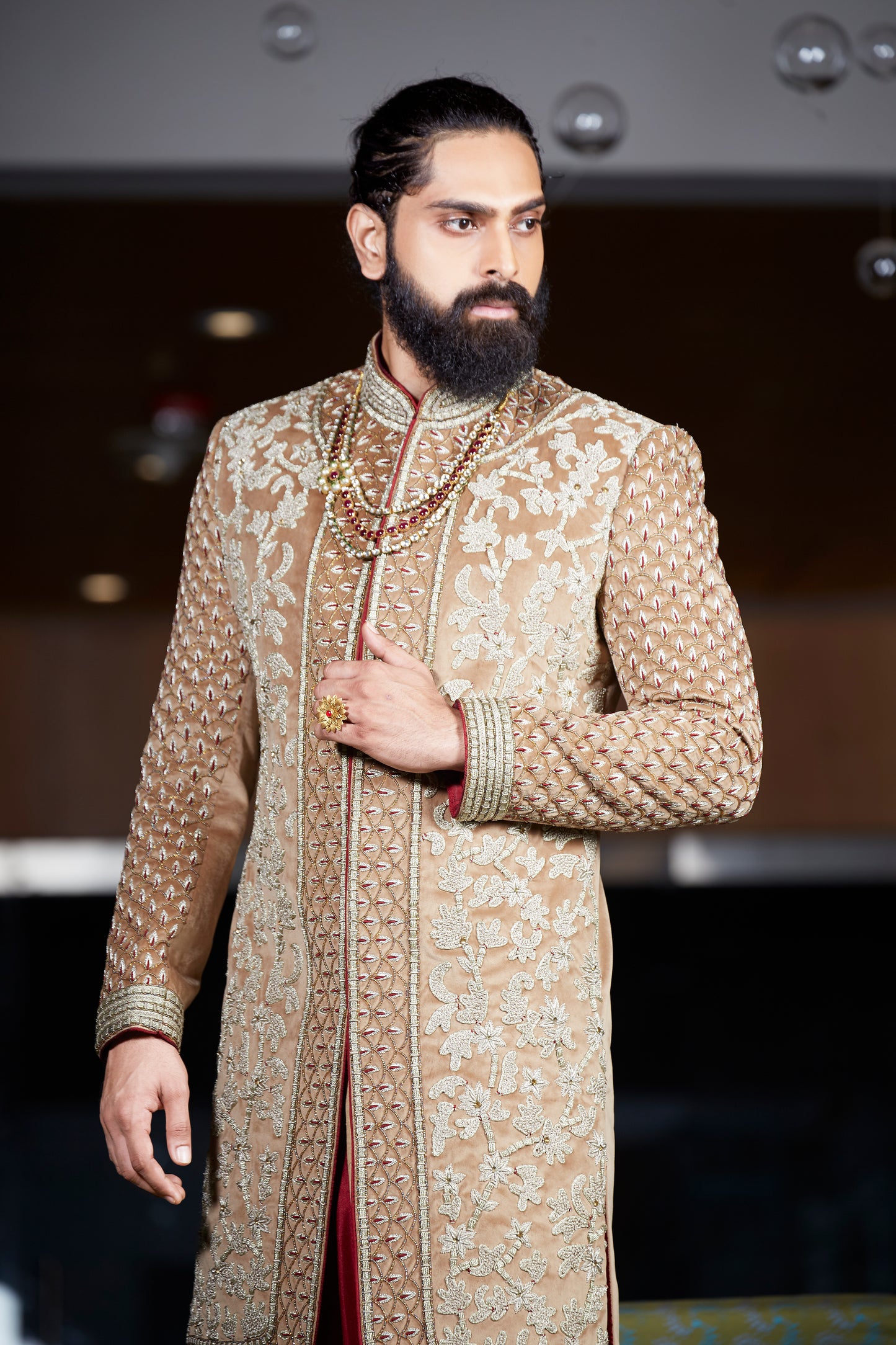 A man with a beard and styled hair wearing a beige and gold heavily embroidered sherwani jacket, standing in a dimly lit indoor setting.