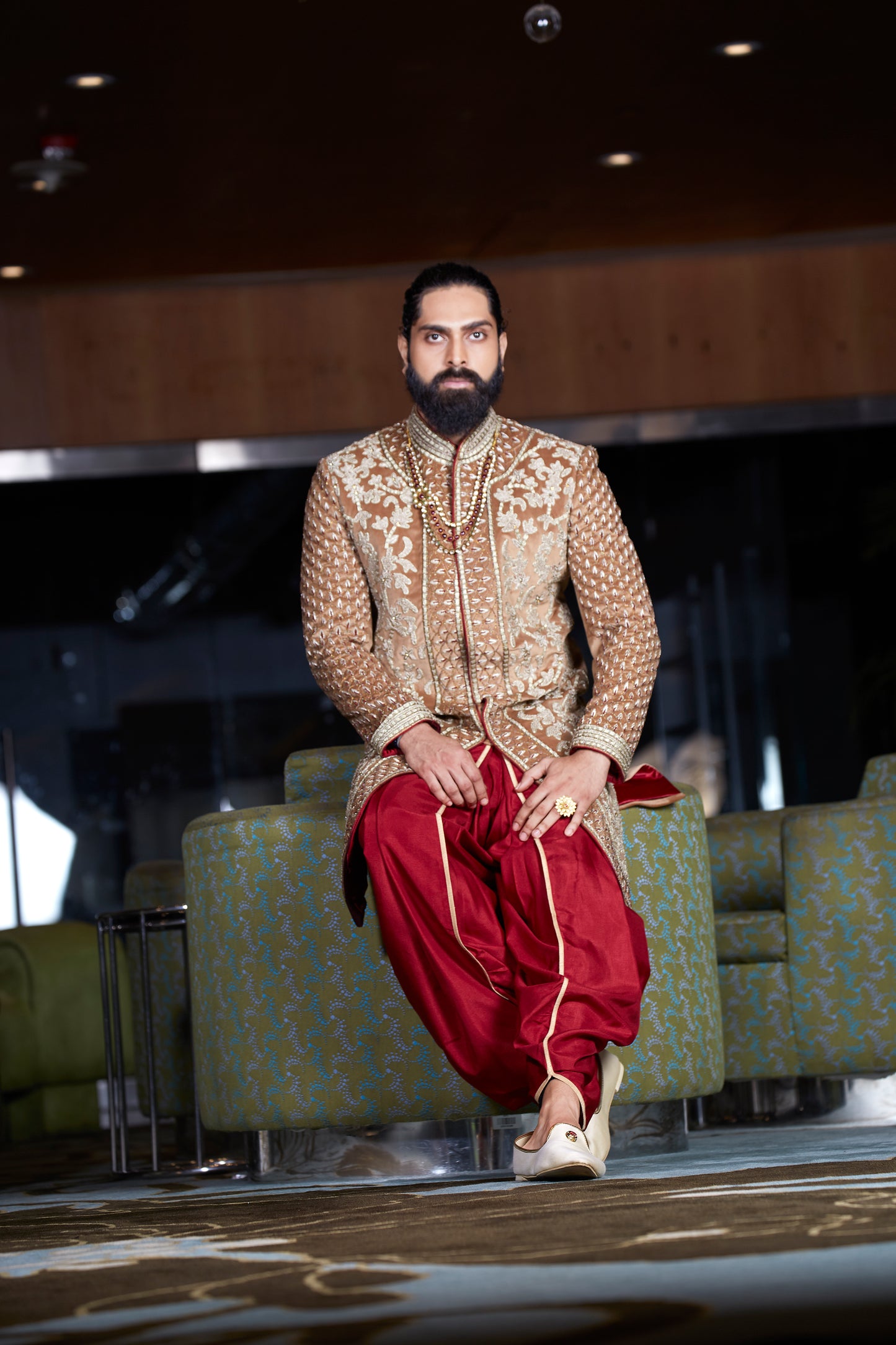 A man wearing a traditional Indian outfit consisting of a beige embroidered sherwani and red patola pants, sitting on a patterned green sofa in a modern indoor setting.