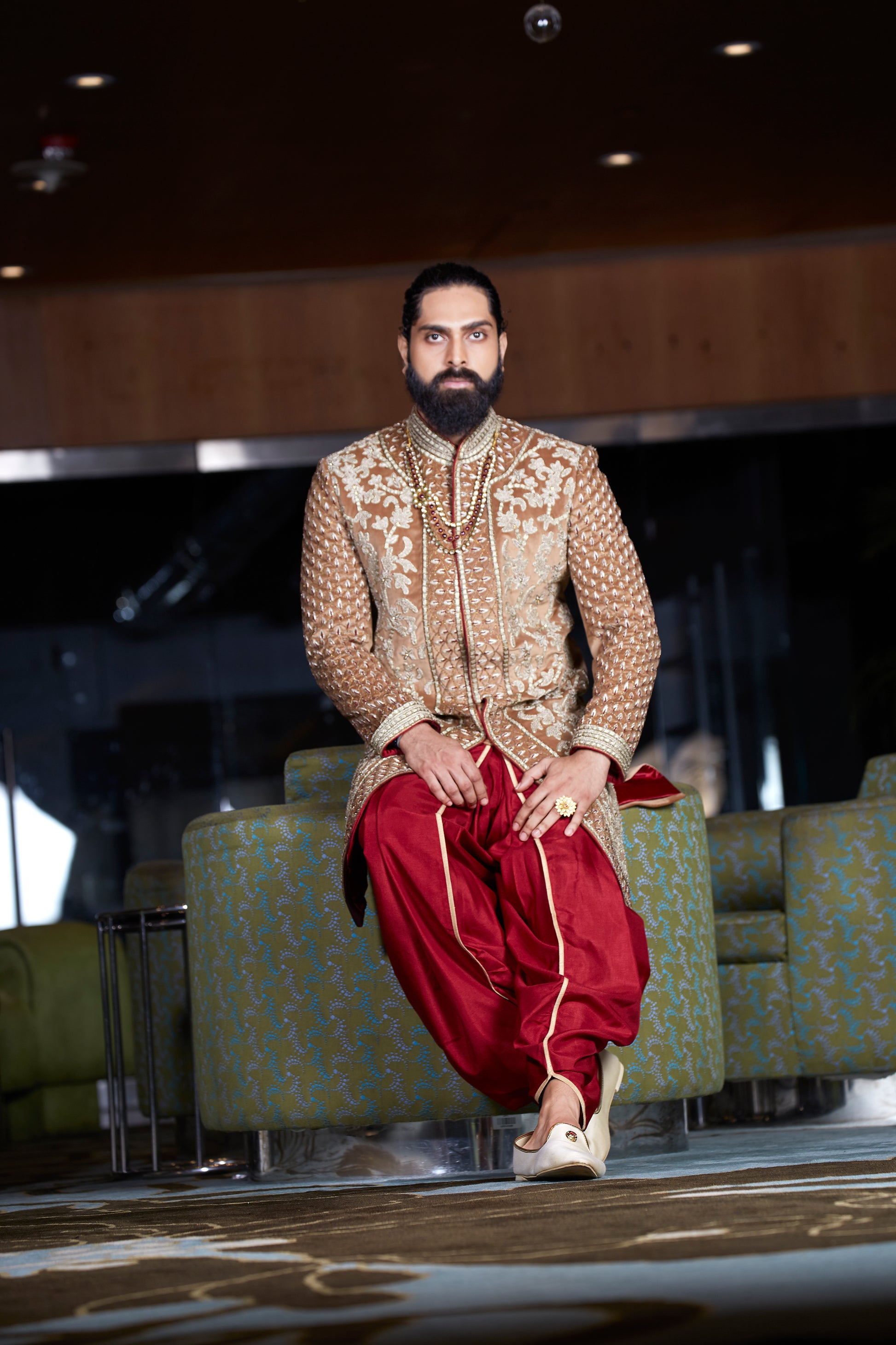 A man wearing a traditional Indian outfit consisting of a beige embroidered sherwani and red patola pants, sitting on a patterned green sofa in a modern indoor setting.