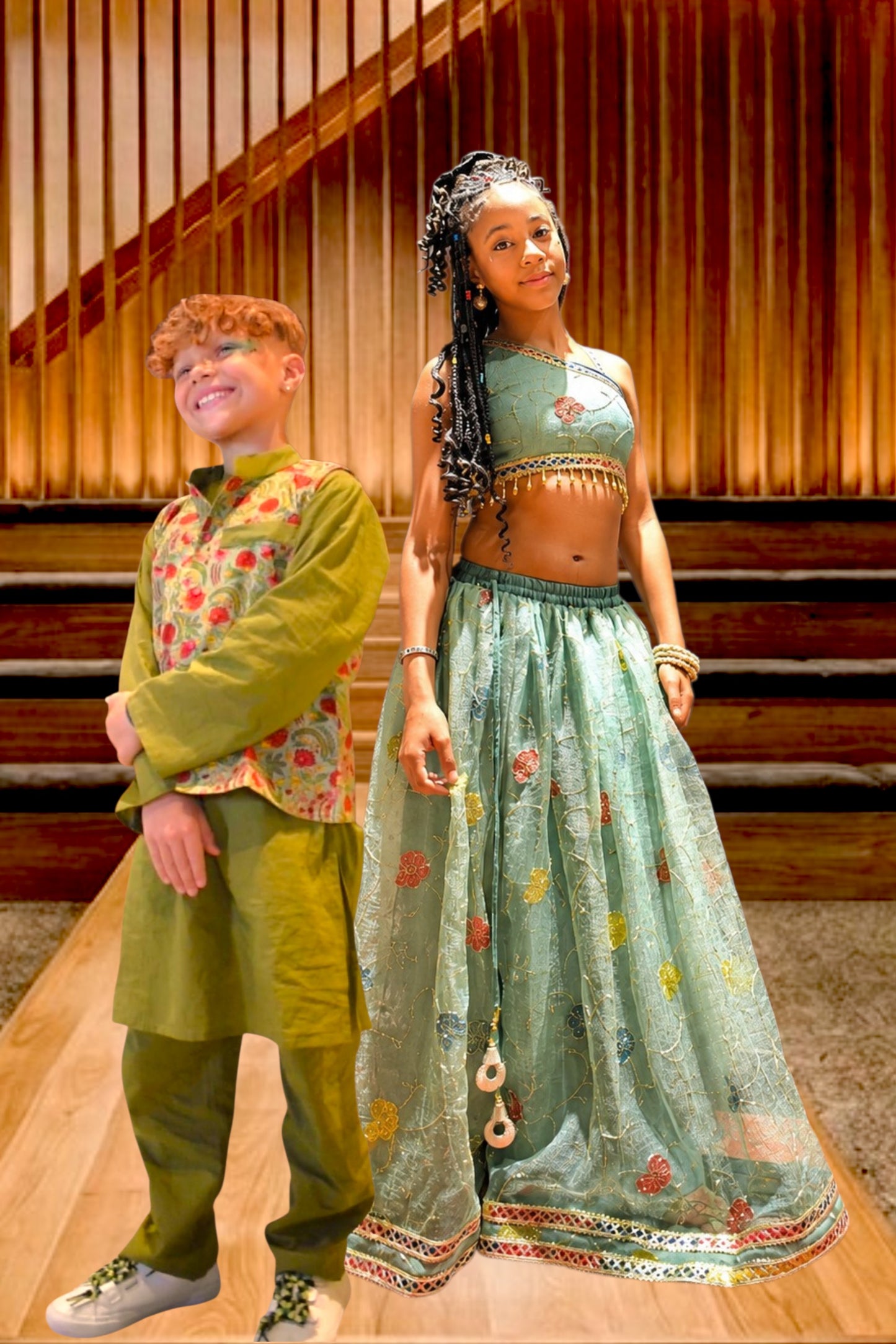 Two children modeling traditional Indian ethnic wear, a boy in an olive green kurta set with a floral embroidered vest and a girl in a mint green lehenga choli with gold embellishments, posed in front of a wooden staircase background.