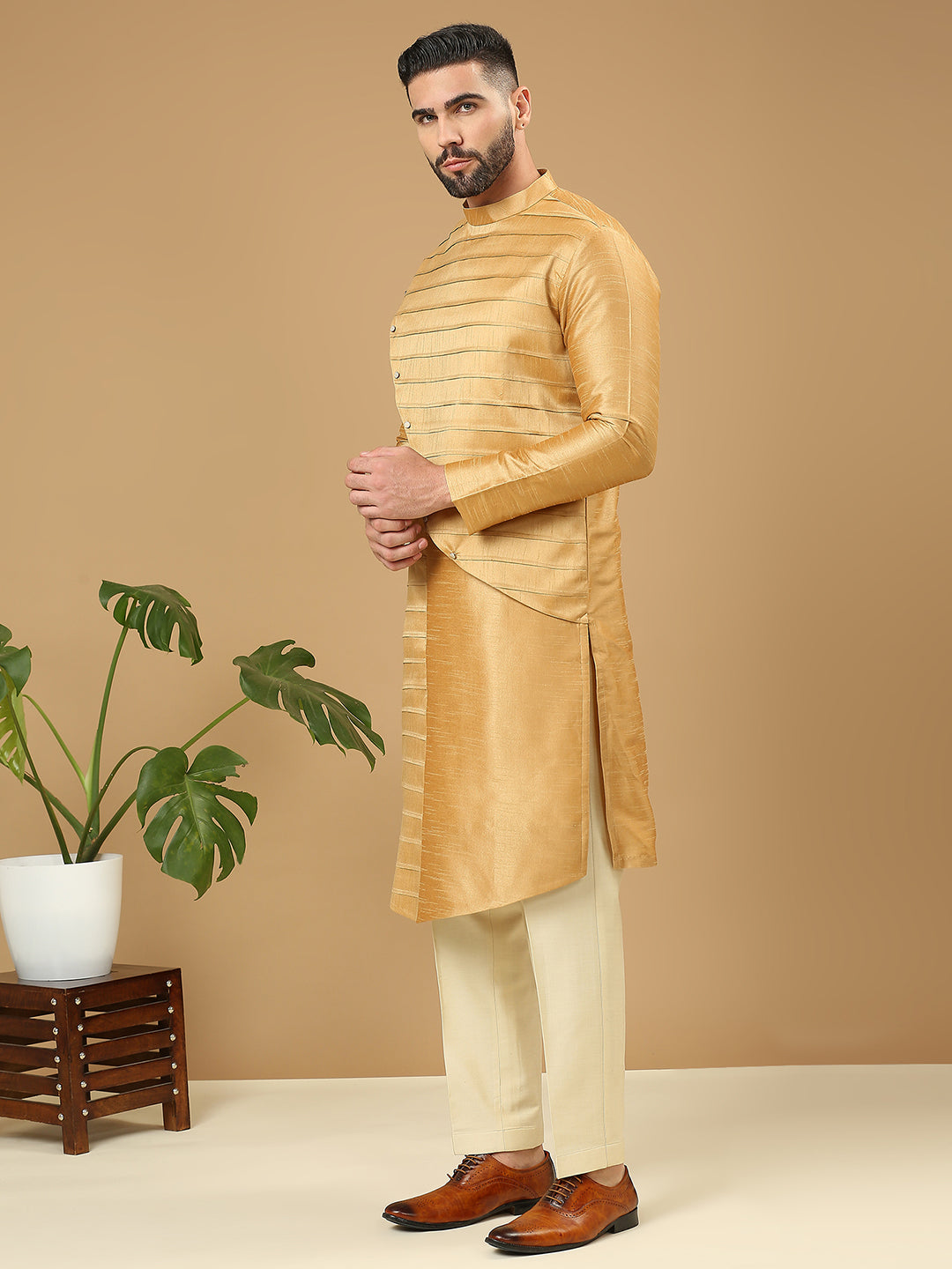 A man modeling a gold long-sleeve kurta with horizontal stripes, paired with cream trousers and brown dress shoes, standing against a solid beige background next to a potted plant.