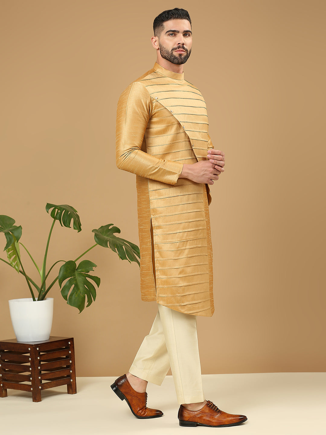 A man modeling a golden-yellow striped kurta with cream trousers and brown leather shoes, standing against a solid beige background next to a potted plant.