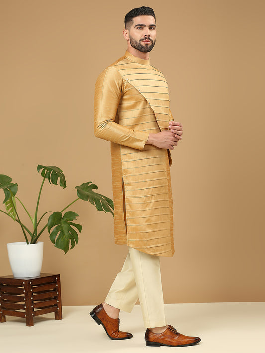 A man modeling a golden-yellow striped kurta with cream trousers and brown leather shoes, standing against a solid beige background next to a potted plant.