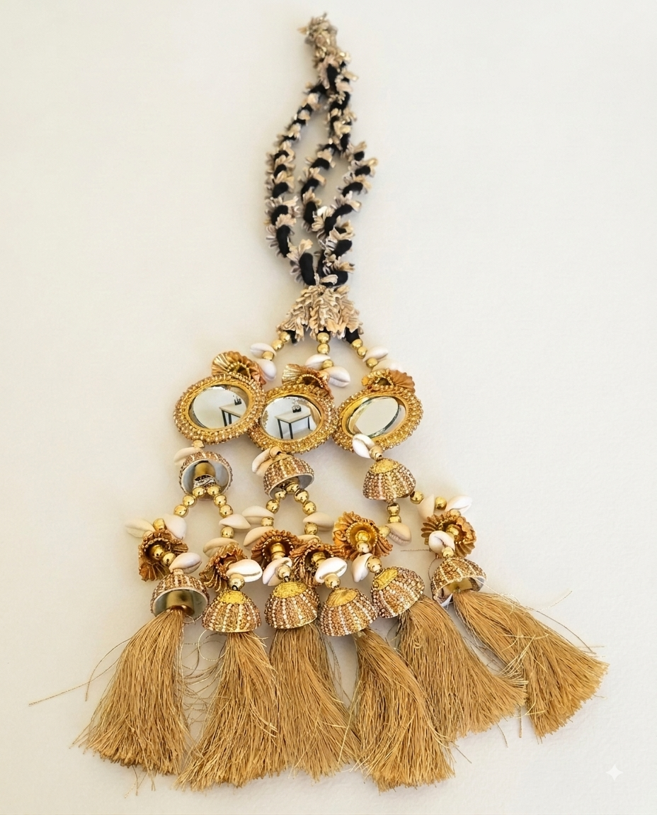 A decorative tassel ornament with gold-toned metalwork, small oval mirrors, white beads, and multiple tan thread tassels hanging from a black-and-gold braided top, shown against a plain light background.