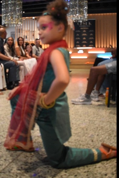 A young child wearing a teal traditional outfit with a red scarf poses on a stage in front of an audience. The child has pink face paint and gold bangles.