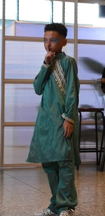 A young boy wearing a traditional green kurta and matching trousers with gold embroidery, standing in an indoor setting with a windowed wall behind him.