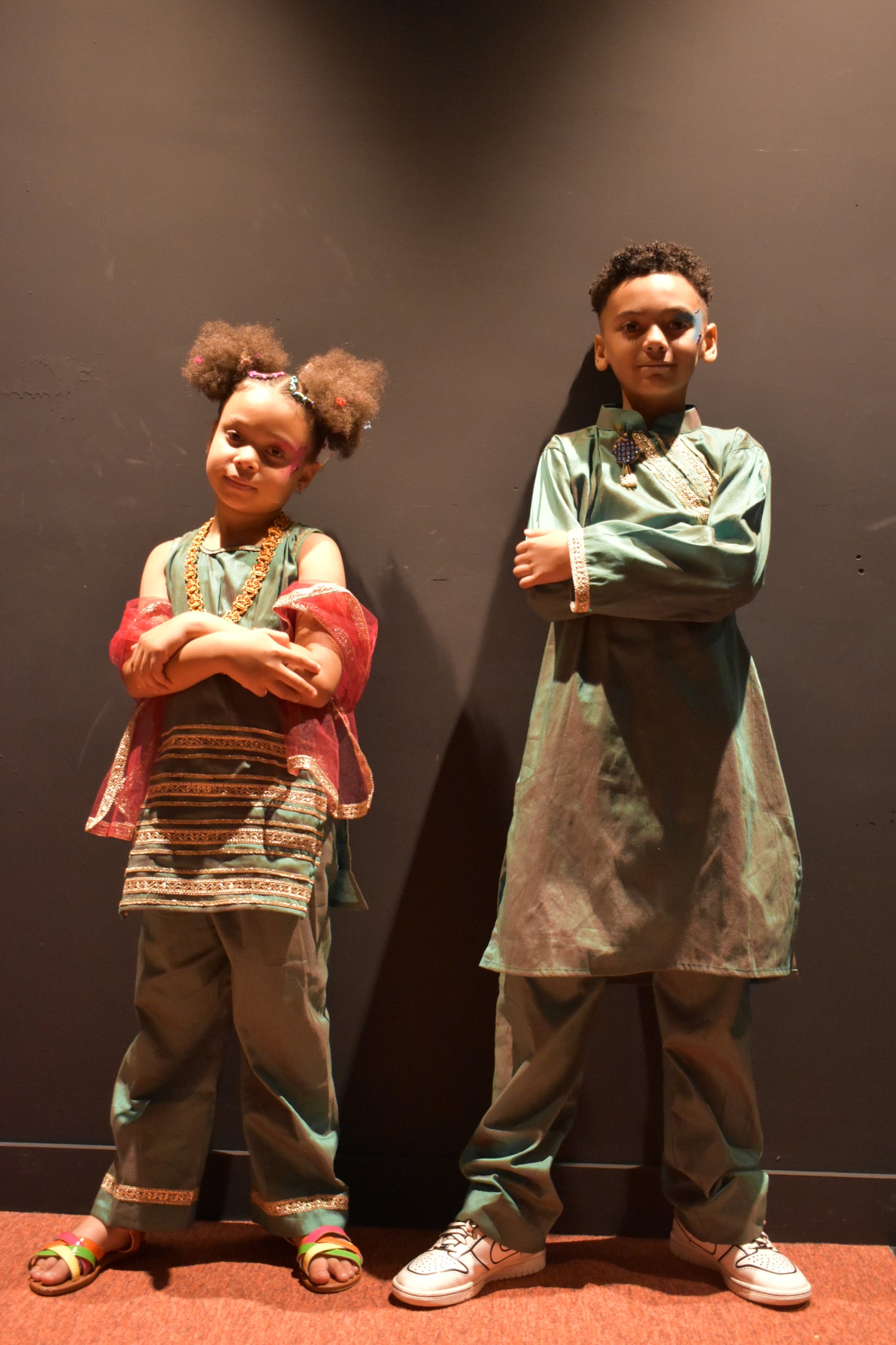 Two children modeling traditional South Asian clothing, standing against a dark wall. The child on the left wears a patterned green and red kurta with matching pants, and the child on the right wears a light green kurta with gray trousers.