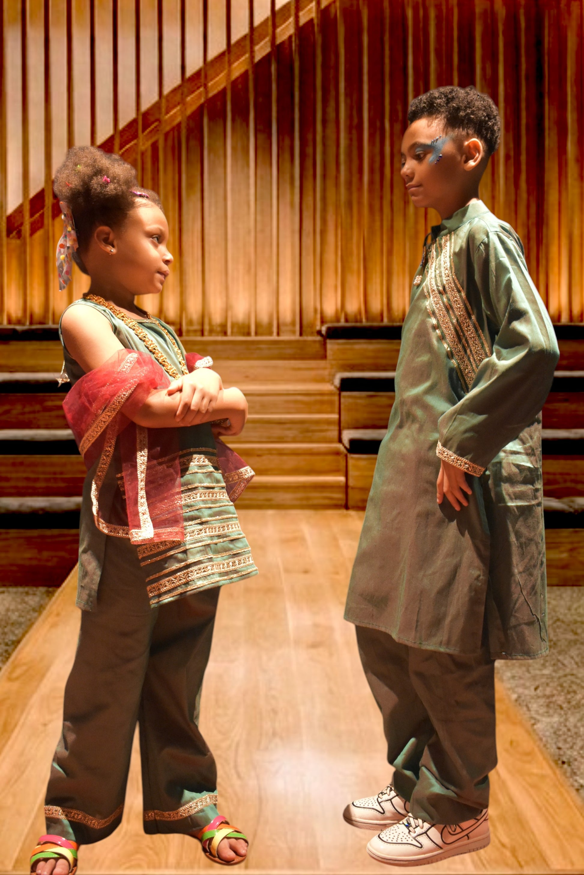 Two children modeling traditional ethnic clothing in a wooden hall. The child on the left wears a colorful off-the-shoulder tunic and pants, while the child on the right wears a long sage green tunic with embroidered details.