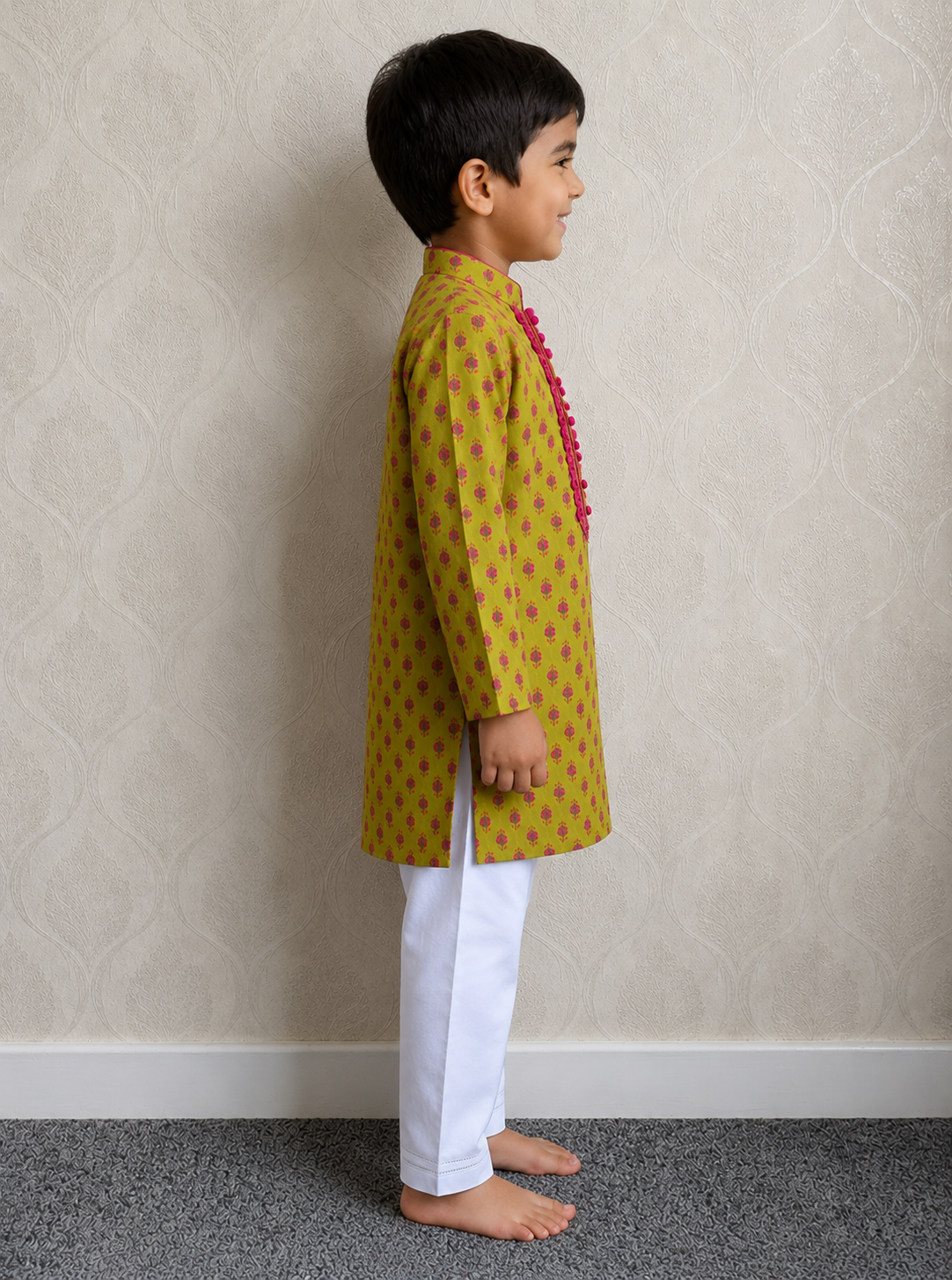 A young boy standing in profile wearing a green kurta with small pink motifs and a pink tassel detail, paired with white trousers, against a textured beige wall.