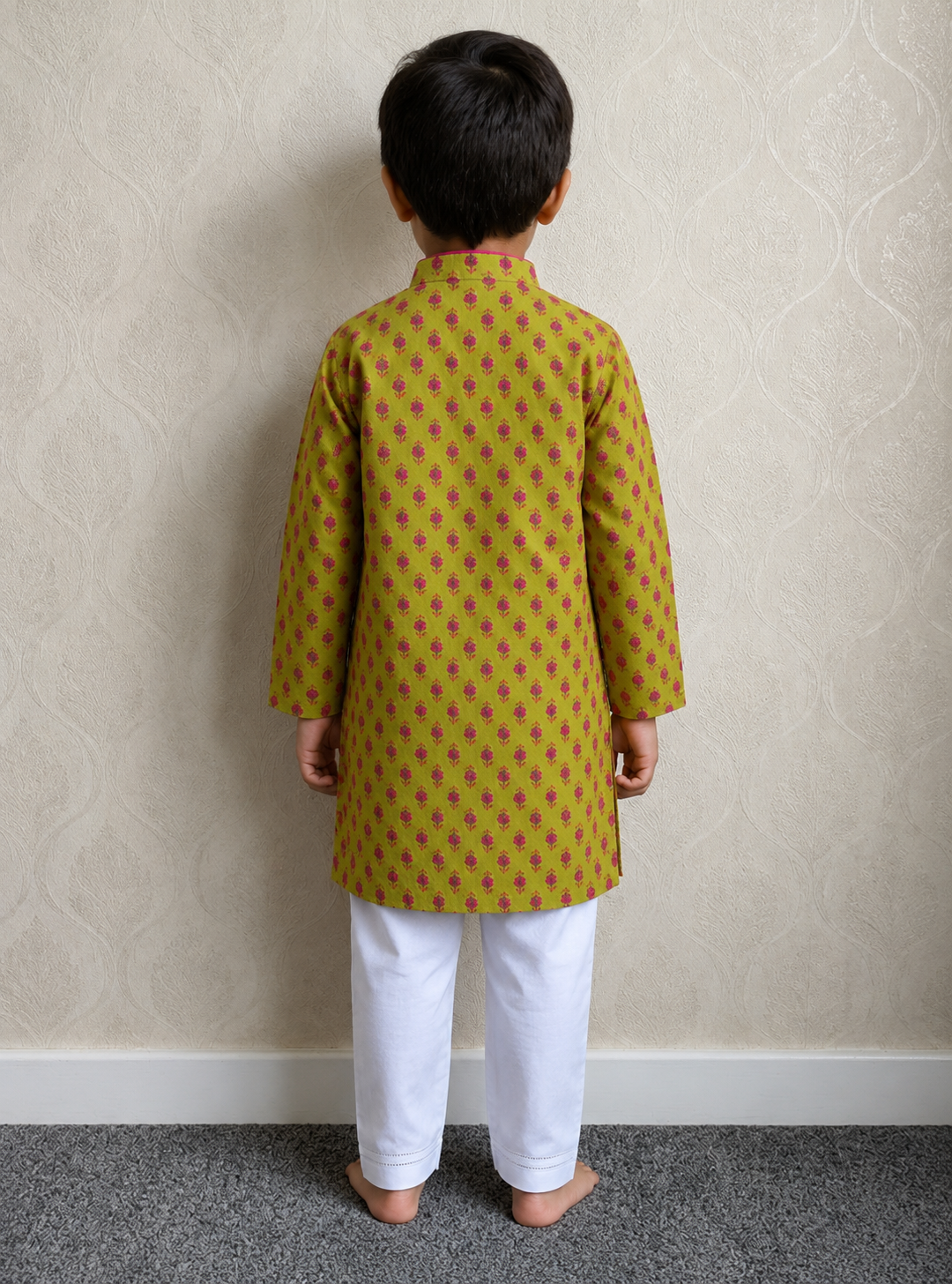 A child standing with their back to the camera, wearing a green kurta with a pink floral pattern and white pants, set against a beige patterned wallpaper background.