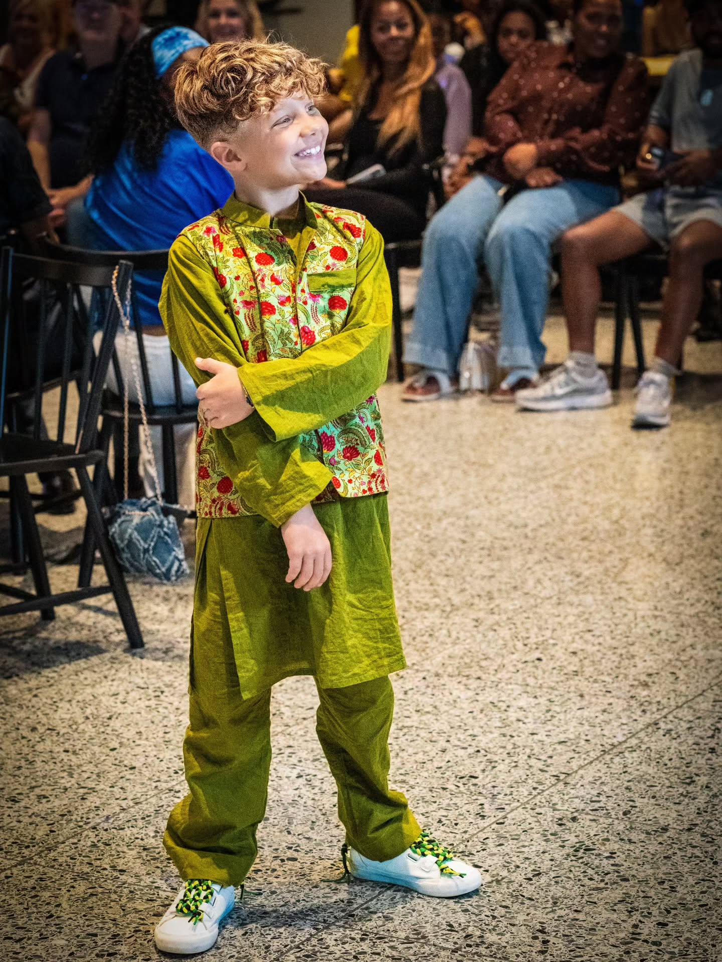 A young boy with curly blond hair stands in a fashion show audience wearing a bright olive green traditional outfit with a floral patterned vest and white sneakers with green details.