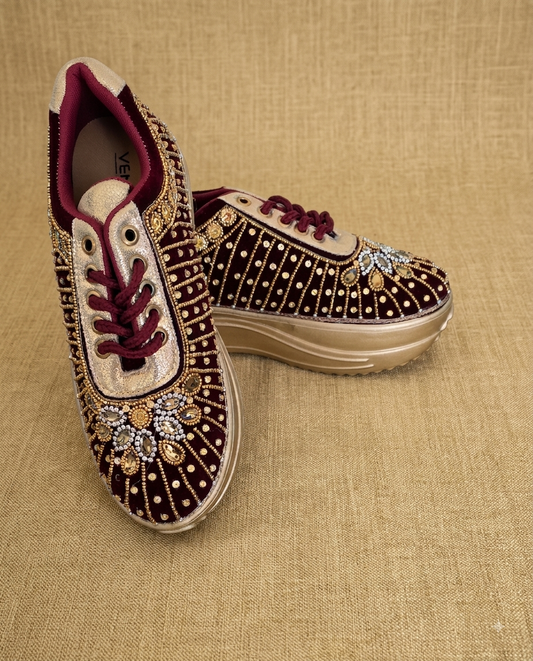 A pair of low-top platform sneakers with deep burgundy velvet uppers heavily embellished with gold and silver rhinestones, featuring burgundy laces and a beige platform sole, set against a textured beige fabric background.