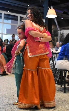 A woman in a vibrant orange and pink traditional Indian outfit hugging a child wearing a teal kurta and pink dupatta in an indoor event space.