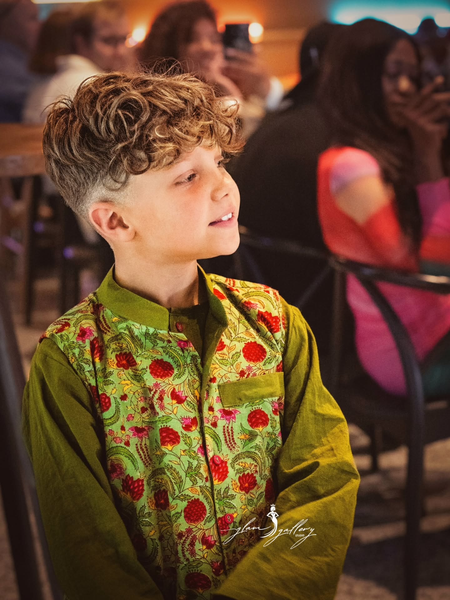 A young boy with curly light brown hair wearing a green embroidered traditional-style jacket, seated in a crowded indoor event space with blurred people in the background.