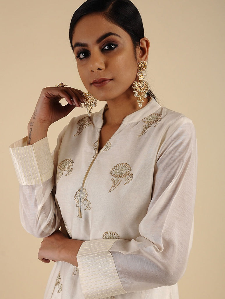 A woman modeling a cream-colored traditional kurta with gold floral embroidery, paired with matching cream trousers and ornate gold earrings, set against a solid beige background.