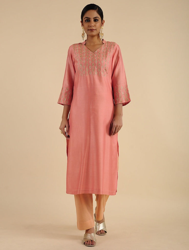 A woman modeling a peach-pink kurta with silver embroidery on the chest and sleeve cuffs, paired with beige pants, against a solid beige background.