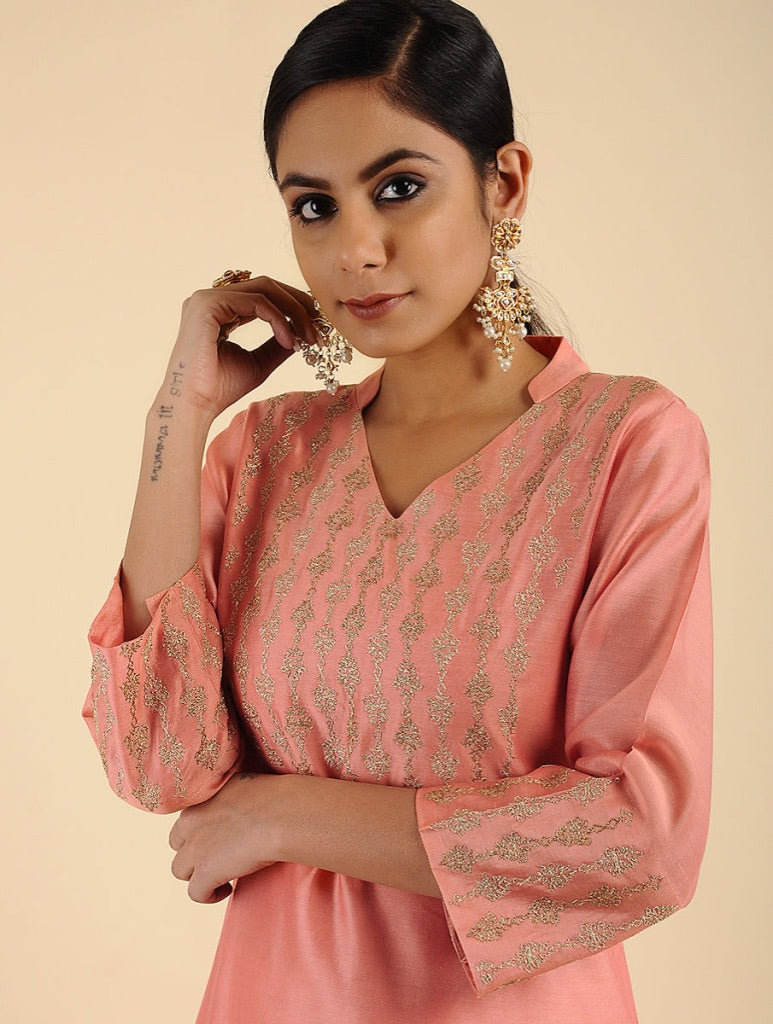 A woman modeling a peach-pink kurta with gold metallic embroidery, featuring a V-neckline and elbow-length sleeves, set against a solid beige background.