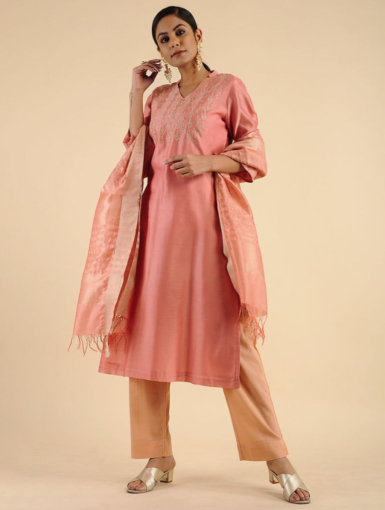 A woman models a peach-pink kurta set with matching trousers and a patterned dupatta against a solid beige studio background.