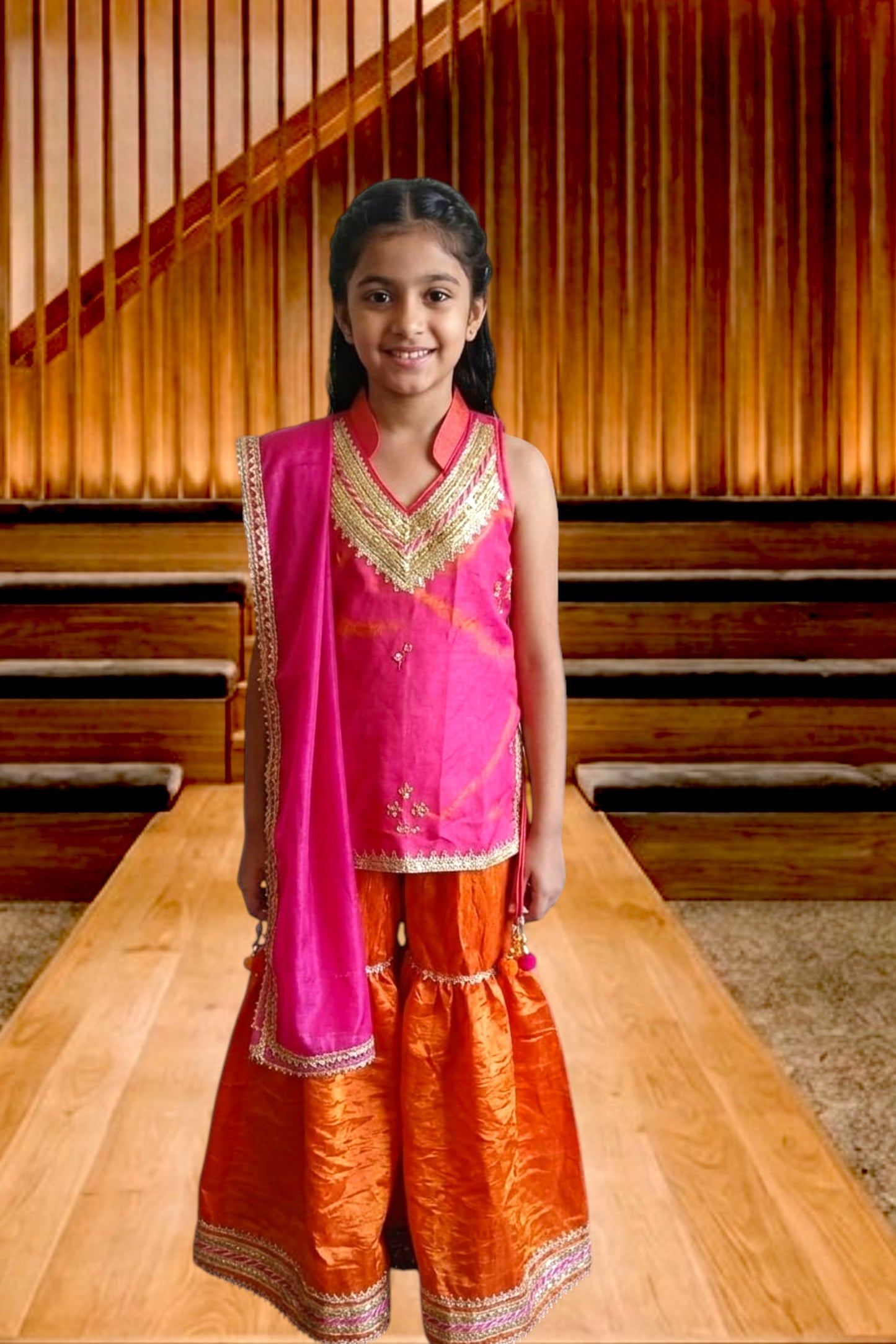 A young girl stands on a wooden runway wearing a traditional Indian outfit consisting of a bright pink sleeveless top with gold embroidery, orange flared pants, and a pink dupatta with gold borders. The background features a wooden slatted wall and steps.