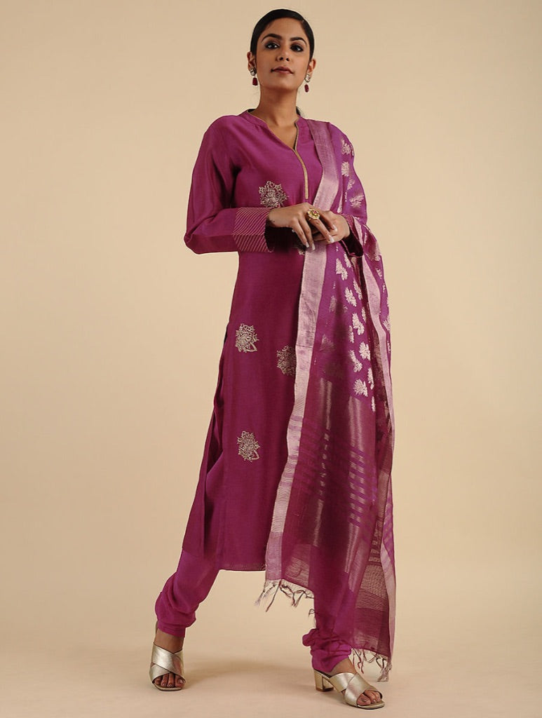 A woman modeling a magenta purple traditional Indian outfit with matching pants and a sheer patterned dupatta, set against a solid beige studio background.