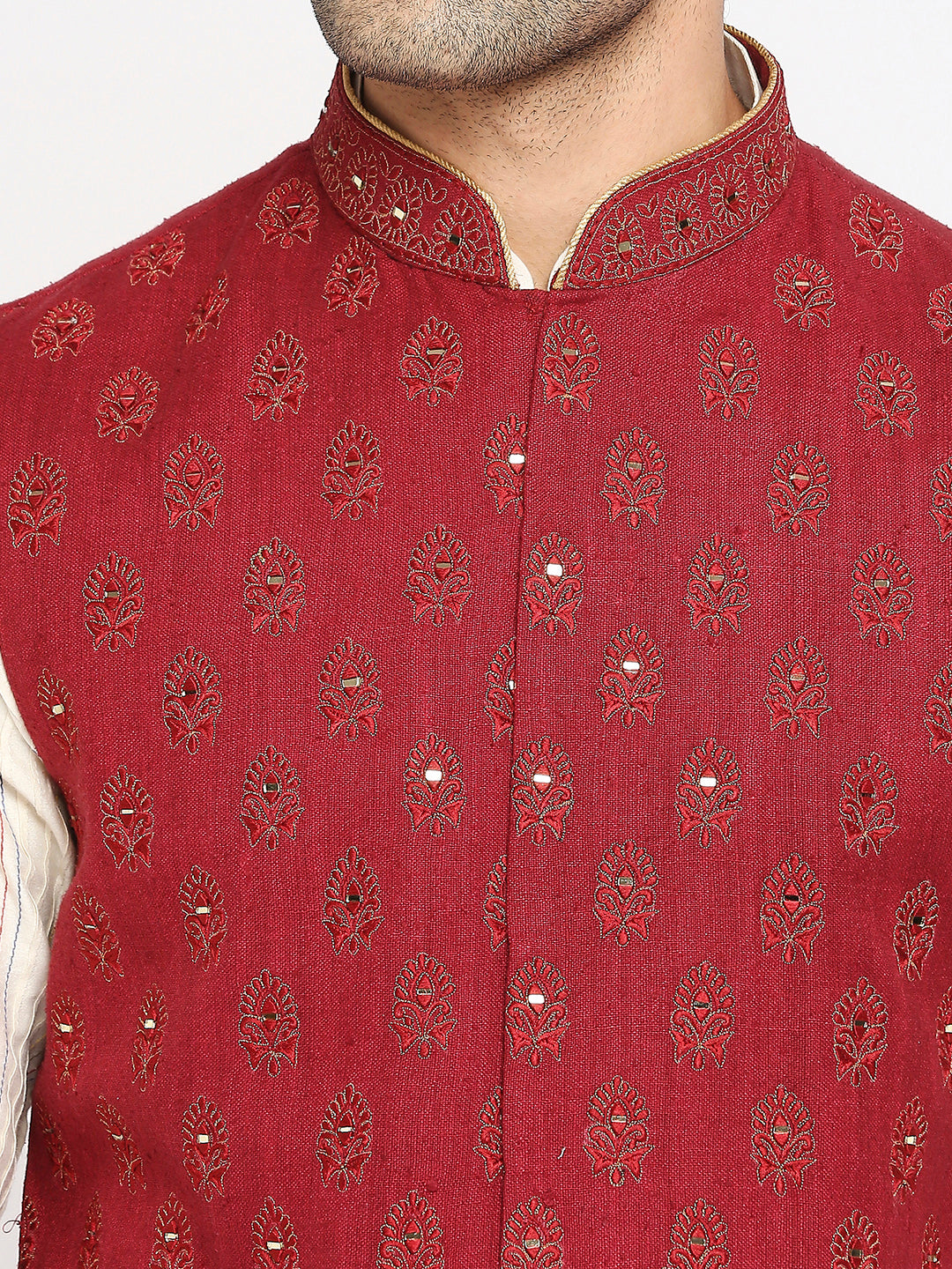 A close-up of a man wearing a red sherwani jacket with gold embroidery and small silver mirror work, paired with a cream shirt.