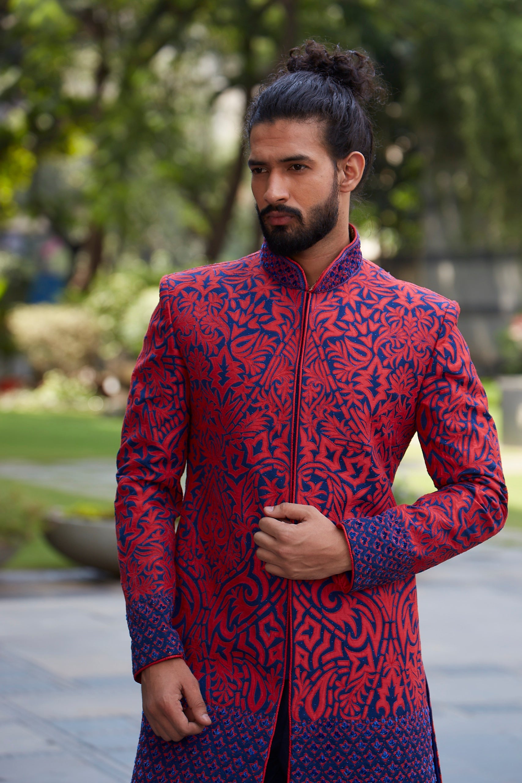 A man wearing a red and navy blue patterned sherwani jacket, standing outdoors in a park-like setting with blurred green trees in the background.