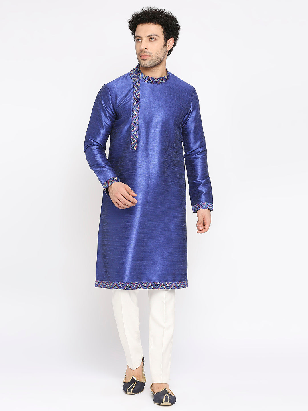 A man modeling a royal blue kurta with gold geometric embroidery on the neckline and hem, paired with white trousers and black-and-gold loafers, against a plain white studio background.