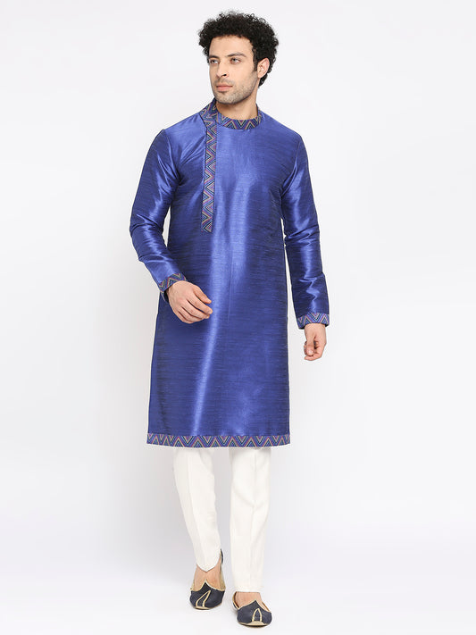 A man modeling a royal blue kurta with gold geometric embroidery on the neckline and hem, paired with white trousers and black-and-gold loafers, against a plain white studio background.