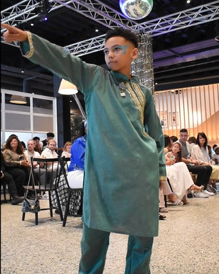 A young boy models a teal green traditional long-sleeve kurta with gold embroidery and matching trousers on a fashion runway, with an audience seated in the background.