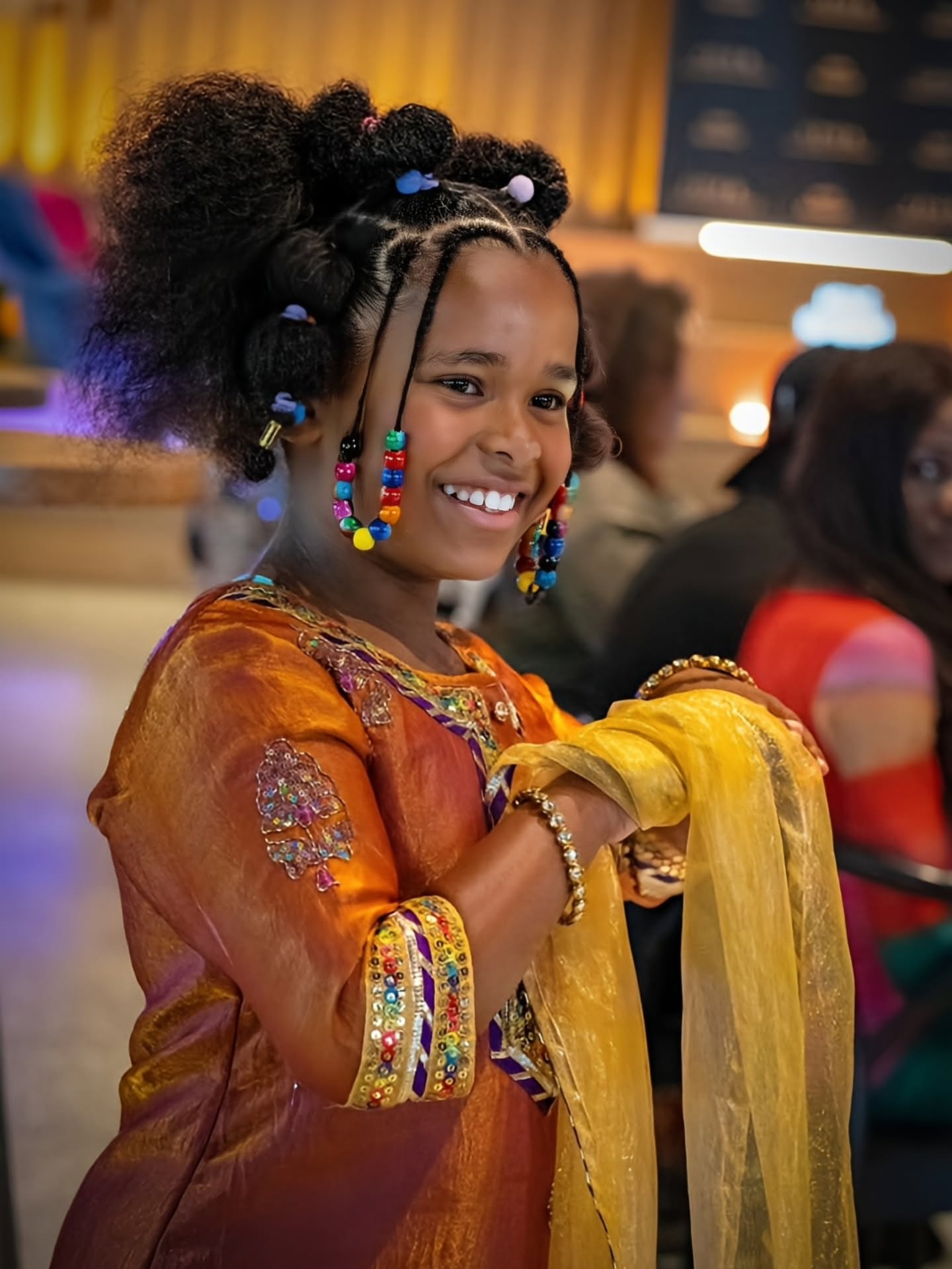 A smiling young girl with elaborate braided hair and colorful beaded earrings, wearing a bright orange embroidered traditional outfit with a yellow sheer scarf, photographed in a warmly lit indoor event setting.