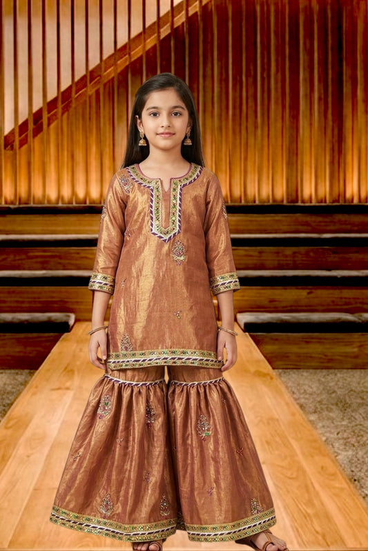 A young girl modeling a traditional brown sharara suit with gold zari embroidery and green accents, standing on a wooden platform in front of a wooden slatted background.